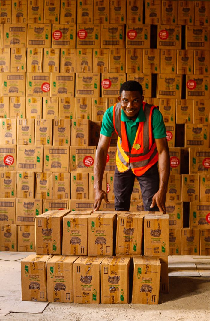 Smiling warehouse worker organizing stacks of boxes with a reflective vest indoors.